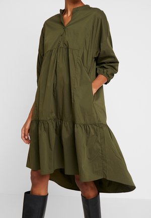 Femme portant une robe ample vert olive à volants froncés, manches longues, encolure sans col et bottes noires montant jusqu'aux genoux.