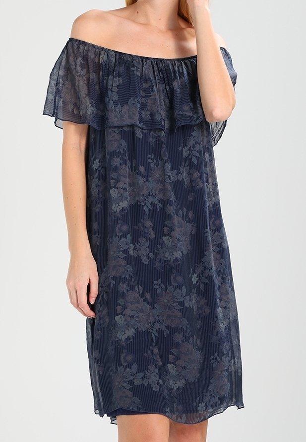 Femme portant une robe bleu marine épaules dénudées avec un motif floral et un col à volants, debout devant un fond clair uni.
