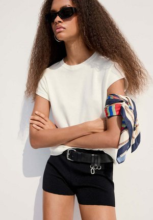 Jeune femme aux longs cheveux bouclés portant des lunettes de soleil noires, un haut blanc à manches courtes, un short noir avec une ceinture et une écharpe colorée nouée autour du bras.