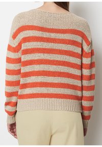 Kobieta w beżowym i pomarańczowym swetrze w paski oglądana z tyłu, w połączeniu z jasnobeżowymi spodniami.