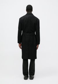 Fekete gyapjú kabát övvel, hosszú ujjakkal és egyenes szabással, sötét nadrággal és fekete cipővel viselve. Hátsó nézet.