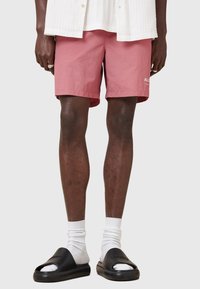Lyserøde shorts lavet af letvægtsmateriale, med to sidelommer og et lille hvidt logo. Båret med sorte klipklapper og hvide sokker.