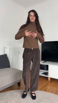 Pull à fermeture éclair marron en maille avec poignets côtelés, accompagné d'un pantalon large en velours côtelé marron foncé et de chaussures noires. Debout sur un tapis clair.