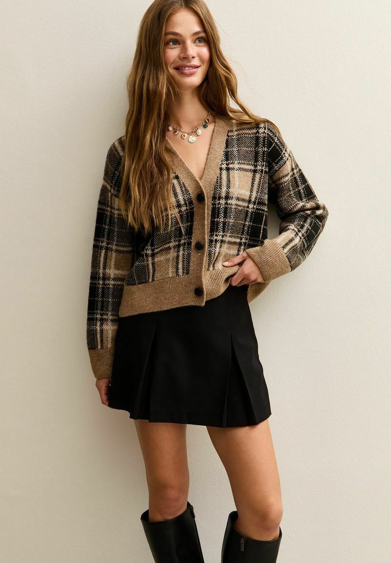 Cardigan à carreaux brun et noir avec un col en V, des poignets côtelés et six boutons. Associé à une jupe mini plissée noire et des bottes jusqu'aux genoux.
