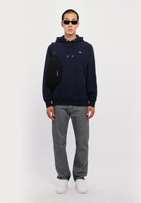 Jūras spēku kapuce, pelēki džinsi un baltas kedas. Modelis valkā melnus saulesbrilles un melnu krustenisku somu ar vienkāršu, gludu dizainu.