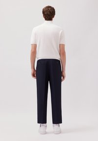 Mann in einem weißen kurzärmeligen Rollkragenpullover, marineblauen Hosen, weißen Socken und weißen Sneakers, der auf einem einfarbigen Hintergrund mit dem Rücken zur Kamera steht.