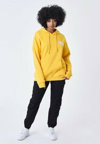 Sudadera amarilla de tela suave con un bolsillo frontal y un gráfico de corazón; combinada con pantalones cargo negros y zapatillas blancas.