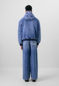 Denim jakna s kapuljačom, plave boje, elastični manžetni. Široke plave traperice s detaljima na stražnjem džepu. Tekstuirani materijal, ležeran kroj.