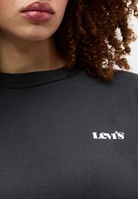 Κοντινό πλάνο ενός ατόμου που φοράει μαύρο πουκά μισο Levi's με καστανά σγουρά μαλλιά ορατά στη δεξιά πλευρά.