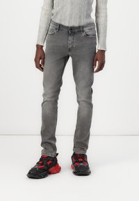 Jeans ajustados grises con textura desgastada, diseño de cinco bolsillos y costuras en contraste, combinados con zapatillas deportivas negras y rojas.