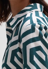 Kortärmad skjorta med ett geometriskt mönster i teal och vitt, tillverkad av lättviktsmaterial och med krage.