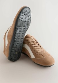 Baskets en daim marron avec des accents blancs, bout rond, et semelle en caoutchouc noir texturé avec un motif en chevrons et un laçage sur le devant.