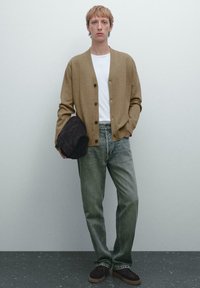 Brun kavaj med knappar, vit t-shirt, avslappnade ljusgröna jeans, bruna mockaskor och en mörk rund hatt. Släta texturer genom hela outfiten.