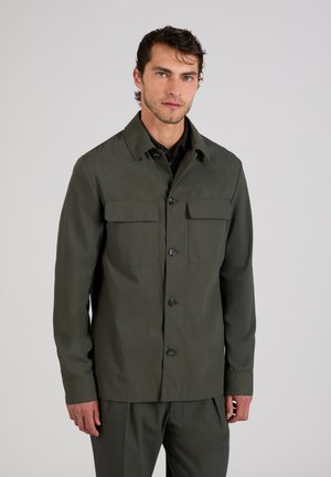 Mężczyzna ubrany w ciemnozieloną kurtkę zapinaną na guziki z klapanymi kieszeniami, stojący na jednolitym jasnoszarym tle.