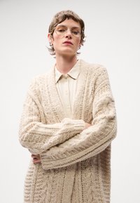 Cardigan beige lavorato a maglia con modelli testurizzati e maniche larghe, abbinato a una camicia button-up di colore chiaro. Il modello sta in piedi con le braccia incrociate.