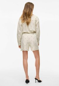 Beżowy zestaw szortów w dwóch częściach z długim rękawem i elastycznym pasem; subtelny geometryczny wzór; noszony z czarnymi wysokimi obcasami.