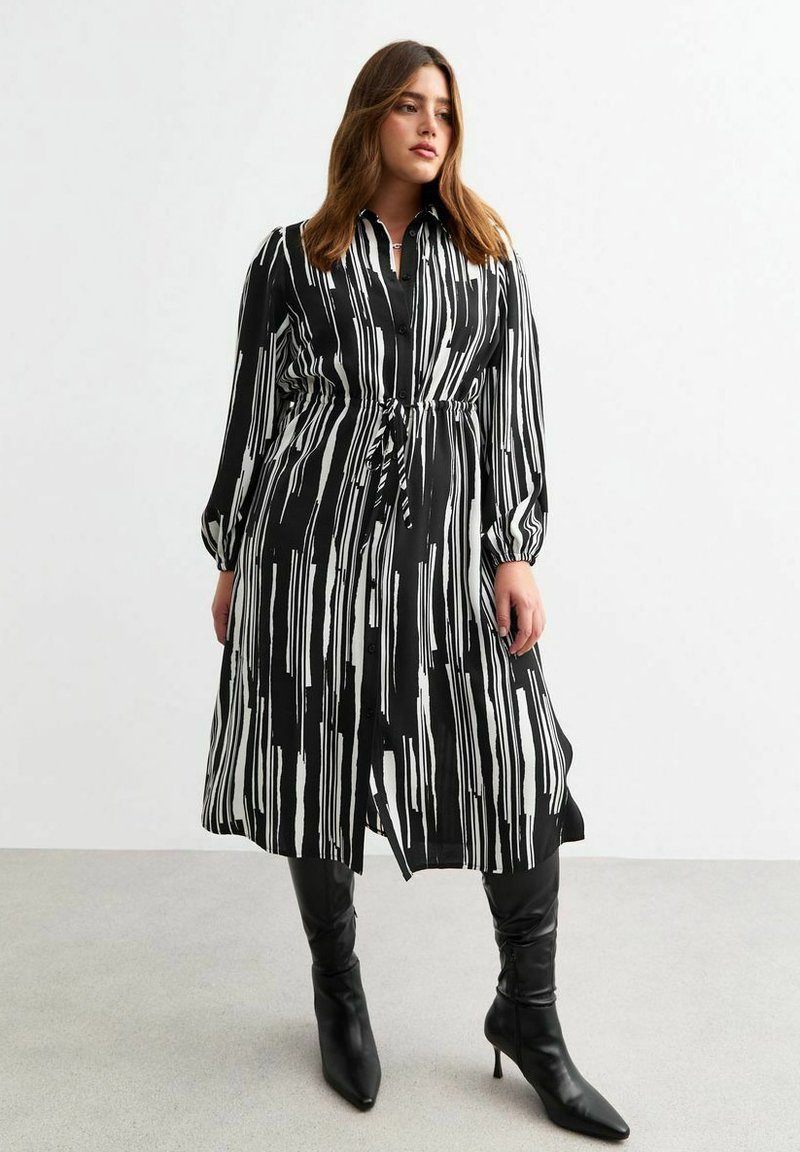 Un lungo vestito a righe bianche e nere realizzato in tessuto leggero, con colletto e cintura da annodare in vita. Abbinato a stivali neri fino al ginocchio.