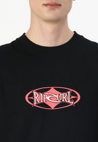 Svart t-skjorte med rund hals og korte ermer. Har en rosa "Rip Curl"-logo i en oval design på fronten. Glatt stofftekstur.