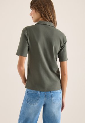 Donna che indossa una polo a maniche corte verde oliva e jeans azzurri, vista di spalle contro uno sfondo neutro.