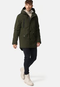 Green parka jacket with hood, black accents, front pockets, drawstring waist, paired with navy pants and light-colored boots.