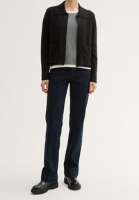 Černá zkrácená bunda s předními kapsami přes kostkovanou košili, doplněná tmavě modrými zvonovými džíny a černými robustními botami.