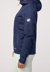 Personne portant une veste softshell à capuche bleu marine avec une poche zippée, un logo sur la manche et un pantalon clair, vue de profil.