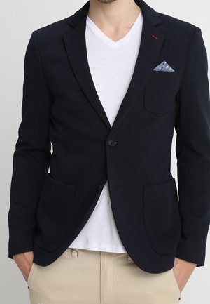 Mand iført navy blazer med et blåt lommetørklæde med prikker, hvid V-hals skjorte og beige bukser med hænderne i lommerne.