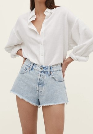 Vrouw met een wit, langärmlig blouse met knopen, ingestopt in lichtblauwe versleten spijkershorts, handen in de zij.