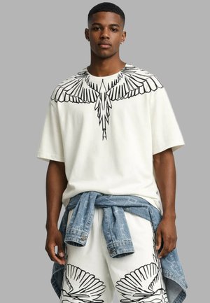 Giovane uomo che indossa una T-shirt bianca oversize e pantaloncini con motivi di ali di uccello nere, giacca di jeans legata in vita, sfondo neutro.