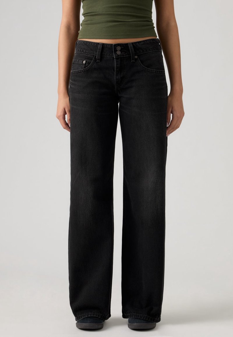 Femme portant un jean noir taille haute à jambes larges et un top court vert olive, debout devant un fond uni.