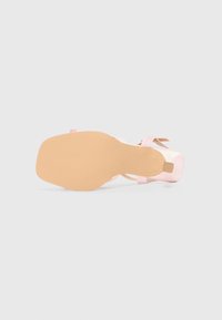 Lyserød højhælet sandal med en glat, blank overflade, firkantet tå og en bred rem dekoreret med tynde, lyserøde ankelbånd. Flad, beige sål.