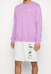 Jasnofioletowa bluza crewneck z miękkiej tkaniny, z białym logo Nike na piersi. Do kompletu białe szorty z wieloma logo.