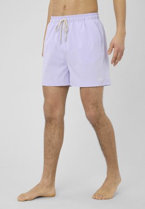 Homme portant un short de bain violet clair avec cordon de serrage, debout pieds nus devant un fond uni.