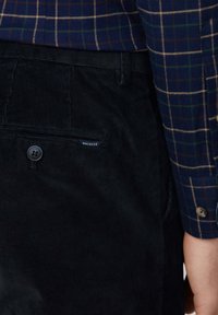 Pantalon en velours côtelé noir avec une poche arrière dotée d'un bouton. Chemise à carreaux bleue accompagnante avec des accents verts et beige.