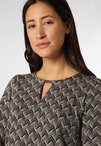 Schwarzes und beiges Oberteil mit Zickzack-Muster, das über einen Schlüssellochausschnitt, lange Ärmel und einen weichen Stoff mit subtiler Textur verfügt.