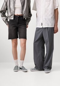 Čierne riflové šortky s rozstrapkaným lemom, kovovo strieborná bomber bunda a šedé široké nohavice v kombinácii so zeleno-bielymi kockovanými slip-on topánkami.