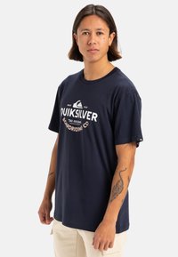 Camiseta de algodón azul marino con un gran logo blanco de Quiksilver, corte relajado, mangas cortas y un pequeño acento del logo en la manga.