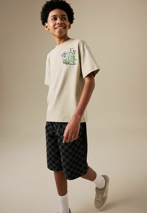 Boy wearing beige graphic t-shirt, black checkered shorts, white socks, and beige sneakers, standing against neutral background smiling.