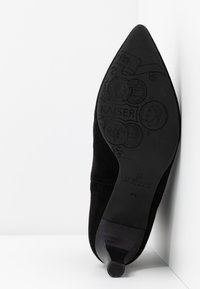 Chaussure à talon haut en daim noir posée sur le côté, montrant la semelle détaillée avec des motifs de pièces en relief et la marque "KAISER". Taille 4½.