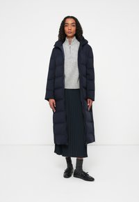 Marineblå lang pufferjakke med hætte, med lynlås foran. Båret over en grå sweater og en plisseret mørk nederdel, parret med sorte sko.