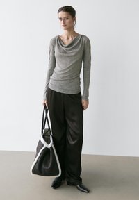 Femme portant un haut gris à col bénitier et manches longues, ainsi qu'un pantalon large noir, tenant un grand sac fourre-tout noir avec une bordure en peau blanche.