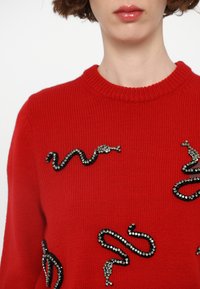 Red knitted sweater featuring black gemstone snake embellishments. Round neckline, textured fabric, and fitted design.