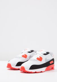 Vita atletiska skor i vitt med svarta och neonorange detaljer, nätöverkärl, rundad tå och synlig luftdämpning i sulan. Lättviktsdesign.