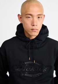 Μαύρη φούτερ με κουκούλα που ρυθμίζεται με κορδόνι, διακοσμημένη με ένα υφασμάτινο λογότυπο σε μαύρο χρώμα στο μπροστινό μέρος. Το υλικό φαίνεται μαλακό, με απαλό υφή.