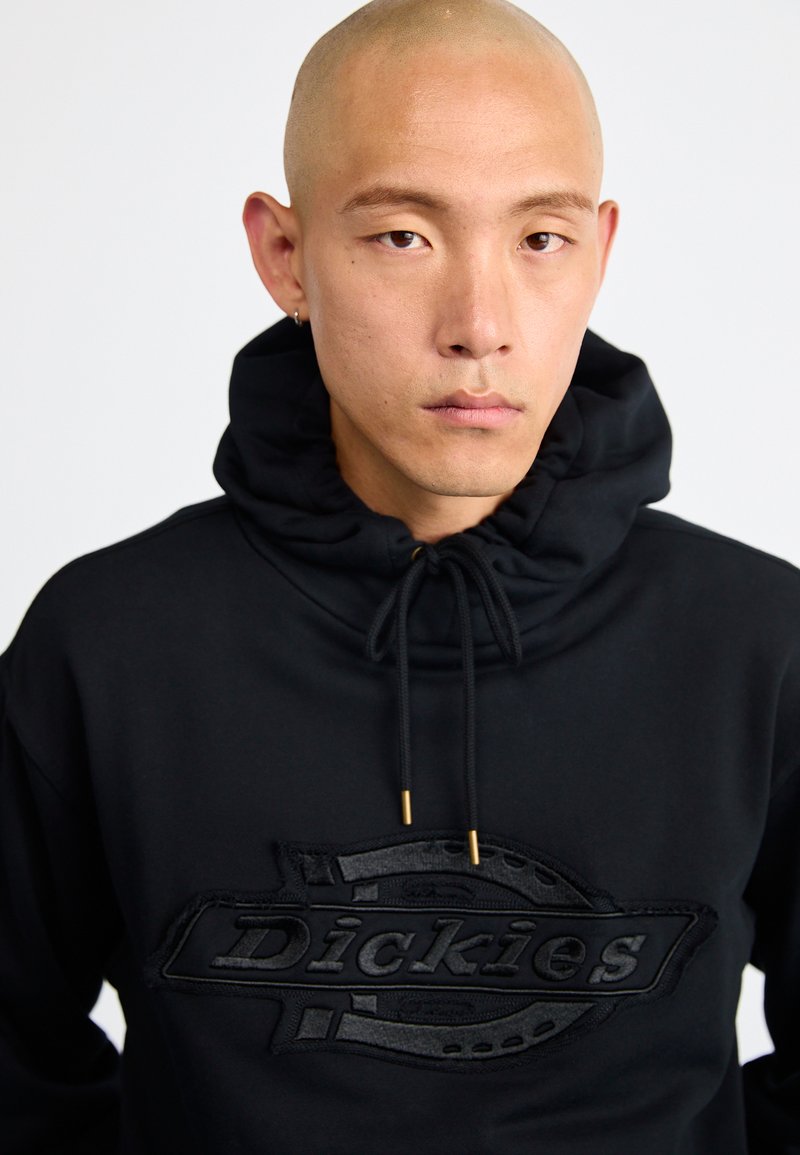 Felpa nera con cappuccio regolabile, caratterizzata da una patch logo nera testurizzata sulla parte anteriore. Il materiale appare morbido, con una texture liscia.