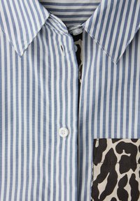 Chemise boutonnée à rayures bleues et blanches, avec une poche imprimée léopard. Confectionnée dans un tissu doux, avec un col classique et un bouton blanc.