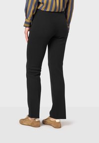 Pantalons noirs ajustés avec une texture lisse et un léger évasement à l'ourlet, associés à des chaussures beiges, vus de dos.