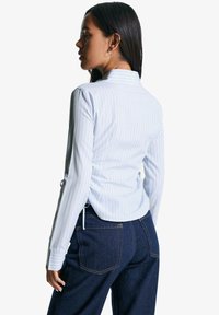 Lichtblauw gestreept overhemd met lange mouwen en een iets geplooid rugdetail, gedragen bij donkere spijkerbroeken.