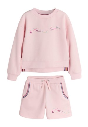 Young girl standing with hands in pockets wearing a pink sweatshirt and matching shorts with white sneakers and socks on a plain background.