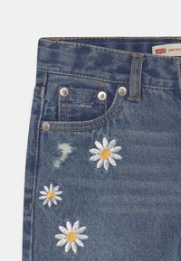 Blå denimjeans med vita och gula broderade prästkragar och slitna detaljer. Inkluderar metallknappar och en klassisk femficksdesign.